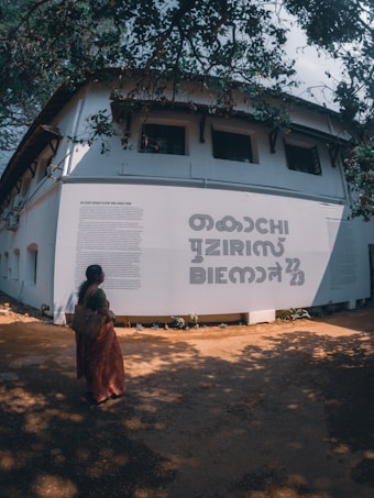 A person in traditional attire stands in front of a large white wall with bold text and smaller text paragraphs. The wall is part of a building with multiple windows, and the scene is shaded by overhanging tree branches and leaves.