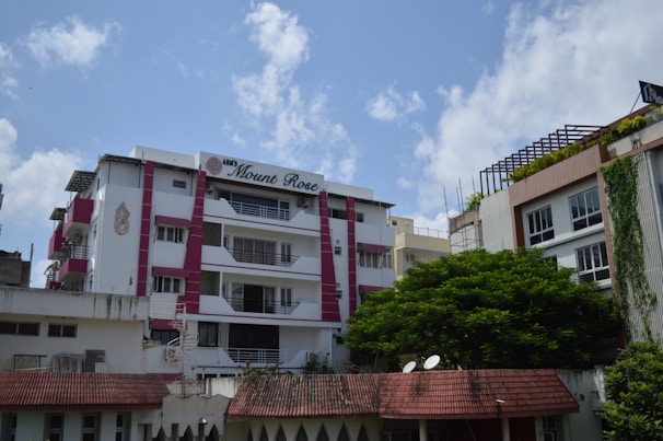 A residential building with multiple floors is visible, featuring a mix of white and pink accents. The sign 'ABM's Mount Rose' is displayed prominently. The building has balconies with railings, and there is a tree with lush green foliage in the foreground. The sky is partly cloudy, providing a bright and airy ambiance.