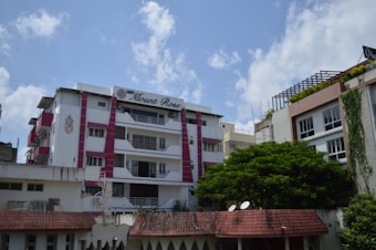 A residential building with multiple floors is visible, featuring a mix of white and pink accents. The sign 'ABM's Mount Rose' is displayed prominently. The building has balconies with railings, and there is a tree with lush green foliage in the foreground. The sky is partly cloudy, providing a bright and airy ambiance.