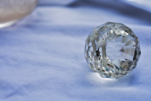 A faceted, transparent crystal orb rests on a soft blue surface, reflecting light through its cuts and edges. The background is out of focus, emphasizing the clarity and pattern of the crystal.