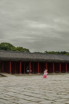 A traditional Korean building with red columns and a tiled roof is situated in the background. The foreground features a large stone courtyard, where a person dressed in a pink hanbok stands, capturing the attention with vibrant traditional attire. The sky is overcast, providing a muted backdrop to the scene.