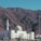 A white mosque with a detailed dome and minaret stands at the base of a rocky mountain. The sky is clear, and the landscape is arid, suggesting a desert environment. Numerous people are gathered around the mosque, wearing traditional garments.