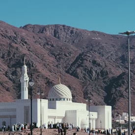 A white mosque with a detailed dome and minaret stands at the base of a rocky mountain. The sky is clear, and the landscape is arid, suggesting a desert environment. Numerous people are gathered around the mosque, wearing traditional garments.