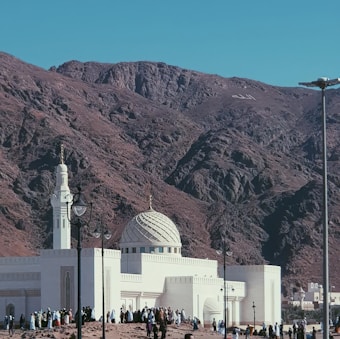 A white mosque with a detailed dome and minaret stands at the base of a rocky mountain. The sky is clear, and the landscape is arid, suggesting a desert environment. Numerous people are gathered around the mosque, wearing traditional garments.