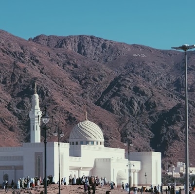 A white mosque with a detailed dome and minaret stands at the base of a rocky mountain. The sky is clear, and the landscape is arid, suggesting a desert environment. Numerous people are gathered around the mosque, wearing traditional garments.