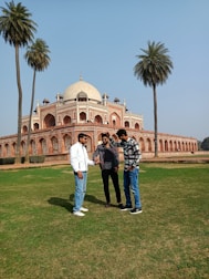 Three people are standing on a grassy area in front of a historical building with distinctive Mughal architecture, featuring a large dome and ornate arches. Tall palm trees are framing the scene, and the sky is clear and bright.