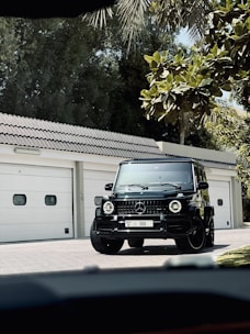 A black Mercedes-Benz G-Class SUV is parked in front of a garage with a tiled roof. The garage doors are white and closed, with a paved driveway leading to them. The background includes lush green trees and some branches hanging down.