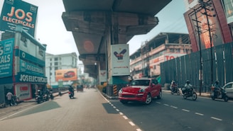 A busy urban street scene featuring a prominent red car on the road with several motorcycles around. Elevated concrete structures loom overhead, possibly part of a transit system, casting shadows on the road. Commercial buildings line the street, including a store called JOS Electricals with vivid signage. Various billboards and advertisements are visible, contributing to the lively city atmosphere.
