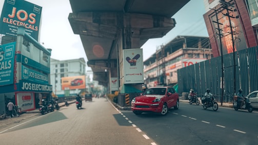 A busy urban street scene featuring a prominent red car on the road with several motorcycles around. Elevated concrete structures loom overhead, possibly part of a transit system, casting shadows on the road. Commercial buildings line the street, including a store called JOS Electricals with vivid signage. Various billboards and advertisements are visible, contributing to the lively city atmosphere.