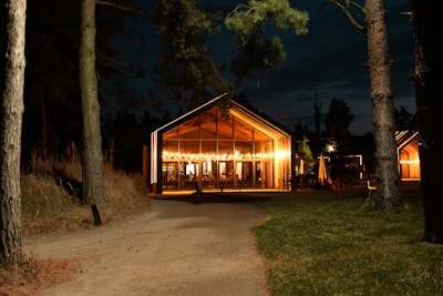A warmly lit wooden structure serves as a cozy gathering space, illuminated against a dark evening backdrop. Surrounded by tall trees, the building emits a welcoming glow, inviting guests inside. The path leads towards the entrance, enhancing the inviting ambiance.