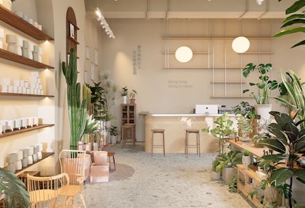 A well-lit, minimalist interior space with natural elements, featuring a variety of green plants and wooden furniture. There are shelves with white ceramic pots on the left, a large cactus, and additional plants scattered throughout. In the back, a wooden counter with stools sits beneath two spherical lamps. The walls are decorated with calming quotes and images, creating a serene and inviting atmosphere.