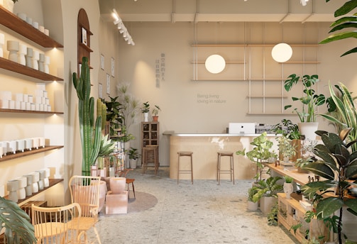 A well-lit, minimalist interior space with natural elements, featuring a variety of green plants and wooden furniture. There are shelves with white ceramic pots on the left, a large cactus, and additional plants scattered throughout. In the back, a wooden counter with stools sits beneath two spherical lamps. The walls are decorated with calming quotes and images, creating a serene and inviting atmosphere.