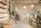 A well-lit, minimalist interior space with natural elements, featuring a variety of green plants and wooden furniture. There are shelves with white ceramic pots on the left, a large cactus, and additional plants scattered throughout. In the back, a wooden counter with stools sits beneath two spherical lamps. The walls are decorated with calming quotes and images, creating a serene and inviting atmosphere.