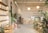 A well-lit, minimalist interior space with natural elements, featuring a variety of green plants and wooden furniture. There are shelves with white ceramic pots on the left, a large cactus, and additional plants scattered throughout. In the back, a wooden counter with stools sits beneath two spherical lamps. The walls are decorated with calming quotes and images, creating a serene and inviting atmosphere.