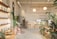 A well-lit, minimalist interior space with natural elements, featuring a variety of green plants and wooden furniture. There are shelves with white ceramic pots on the left, a large cactus, and additional plants scattered throughout. In the back, a wooden counter with stools sits beneath two spherical lamps. The walls are decorated with calming quotes and images, creating a serene and inviting atmosphere.
