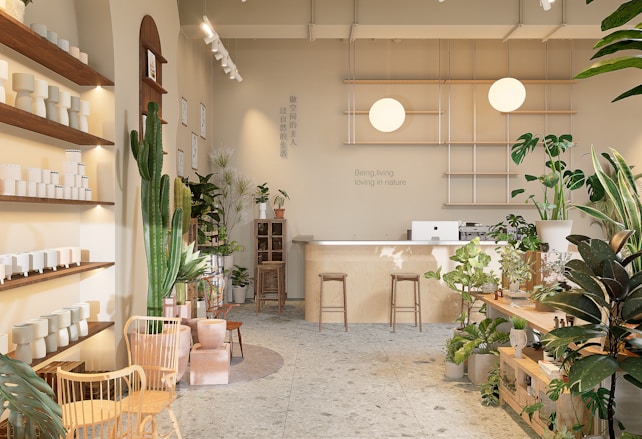 A well-lit, minimalist interior space with natural elements, featuring a variety of green plants and wooden furniture. There are shelves with white ceramic pots on the left, a large cactus, and additional plants scattered throughout. In the back, a wooden counter with stools sits beneath two spherical lamps. The walls are decorated with calming quotes and images, creating a serene and inviting atmosphere.
