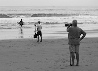 Three people are on a beach with two of them using cameras. One person with a surfboard walks towards the ocean while another is filming with a tripod. A third person stands with a telephoto lens, capturing the scene.