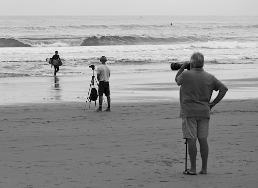 Three people are on a beach with two of them using cameras. One person with a surfboard walks towards the ocean while another is filming with a tripod. A third person stands with a telephoto lens, capturing the scene.