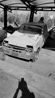 A damaged, old car with a battered front end is parked under a corrugated metal roof. The surroundings include some foliage in the background and an open area with visible paving. The shadow of a person taking a photograph is cast on the ground in the foreground.