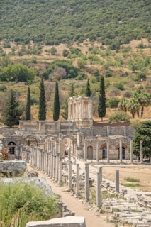 Ancient ruins featuring a row of stone columns leading to a historical structure with multiple arches and fragmented walls. In the background, a hillside densely covered with trees and greenery adds a natural setting. Cypress trees are interspersed around the ruins, contributing to the historical ambiance. The scene suggests a warm and sunny day with clear skies.