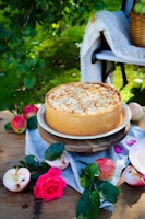 A cheesecake topped with almond slices is placed on a wooden stand outdoors, surrounded by apples, a pink rose, and greenery. A woven basket and a white blanket are on a bench in the background.
