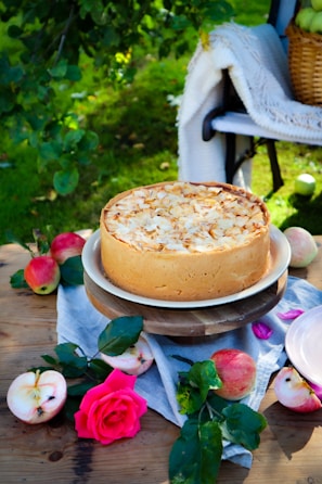 A cheesecake topped with almond slices is placed on a wooden stand outdoors, surrounded by apples, a pink rose, and greenery. A woven basket and a white blanket are on a bench in the background.