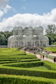 A large glass conservatory structure is situated in the background with lush green gardens and neatly trimmed hedges in the foreground. People are walking along a path that leads to the entrance of the conservatory. The sky above is partly cloudy, providing a picturesque setting.