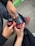 Two hands holding glass Coca-Cola bottles in a casual outdoor setting. One person is wearing red shoes and the scene takes place on a concrete surface, bordered by a patch of green.