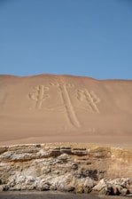 A vast sandy hillside with an ancient geoglyph etched into its surface, resembling a large, stylized candelabra or trident. Below lies a rocky cliff face, with layers of sediment visible, leading to a calm sea at the bottom of the image. The sky above is clear and blue, contrasting with the beige and earthy tones of the desert landscape.