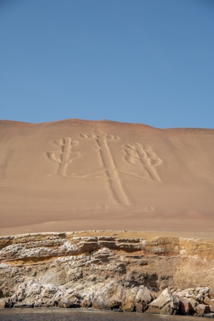 A vast sandy hillside with an ancient geoglyph etched into its surface, resembling a large, stylized candelabra or trident. Below lies a rocky cliff face, with layers of sediment visible, leading to a calm sea at the bottom of the image. The sky above is clear and blue, contrasting with the beige and earthy tones of the desert landscape.