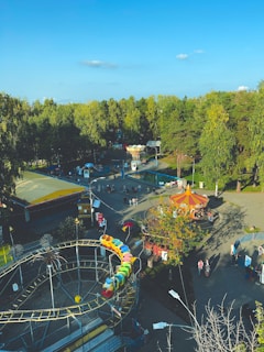 An amusement park scene featuring a colorful caterpillar roller coaster track spiraling towards the foreground. In the background, a circular merry-go-round with a vibrant roof is surrounded by trees. Numerous visitors, including some with strollers, are walking around the area, making it lively.