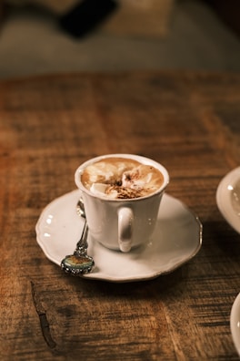 A cup of coffee with frothy milk and sprinkled cocoa on top is placed on a white saucer, with a decorative teaspoon resting on the saucer. The setup is on a rustic wooden table, providing a cozy and warm ambiance.