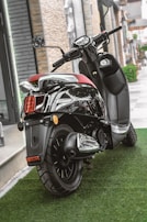 A sleek black scooter parked on a lush green artificial grass surface. The scooter features a shiny chrome exhaust and a comfortable red seat. It is positioned near a modern building with stone and glass elements.