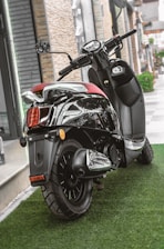 A sleek black scooter parked on a lush green artificial grass surface. The scooter features a shiny chrome exhaust and a comfortable red seat. It is positioned near a modern building with stone and glass elements.