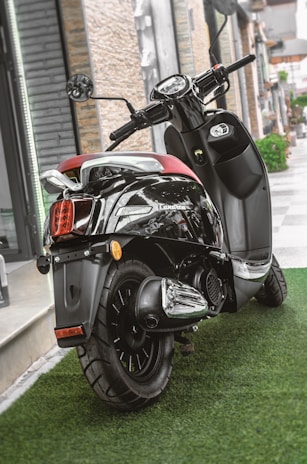 A sleek black scooter parked on a lush green artificial grass surface. The scooter features a shiny chrome exhaust and a comfortable red seat. It is positioned near a modern building with stone and glass elements.