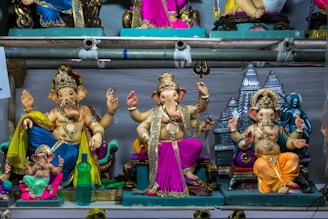 A collection of intricately crafted Ganesh statues in vibrant colors is displayed on shelves. The statues vary in design, with some adorned with decorative embellishments and jewelry. Each figure is posed differently, showcasing a variety of hand gestures and postures typical of Hindu iconography. The background includes temple motifs, adding a spiritual ambiance.