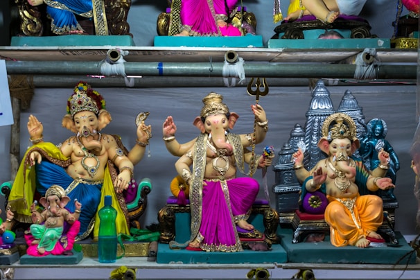 A collection of intricately crafted Ganesh statues in vibrant colors is displayed on shelves. The statues vary in design, with some adorned with decorative embellishments and jewelry. Each figure is posed differently, showcasing a variety of hand gestures and postures typical of Hindu iconography. The background includes temple motifs, adding a spiritual ambiance.