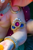 A close-up of a decorative sculpture featuring intricate jewelry designs. The sculpture has a glossy finish with embellishments of pearls and brightly colored stones, including red and blue gemstones. The jewelry includes ornate arm bands and necklaces with gold accents.