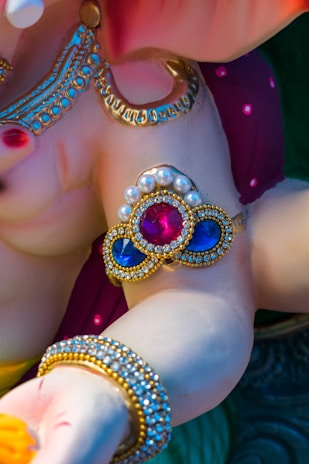 A close-up of a decorative sculpture featuring intricate jewelry designs. The sculpture has a glossy finish with embellishments of pearls and brightly colored stones, including red and blue gemstones. The jewelry includes ornate arm bands and necklaces with gold accents.