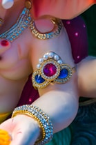 A close-up of a decorative sculpture featuring intricate jewelry designs. The sculpture has a glossy finish with embellishments of pearls and brightly colored stones, including red and blue gemstones. The jewelry includes ornate arm bands and necklaces with gold accents.