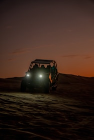 A dune buggy is captured at dusk, with its headlights glowing brightly against a backdrop of dark, rolling sand dunes. The sky is softly lit with an orange gradient, adding a sense of adventure and solitude to the scene.
