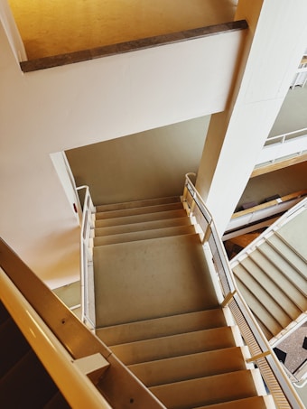 An interior view of a building showing multiple flights of stairs with metal railings. The staircase design features straight lines and geometric patterns, creating a sense of depth and perspective. The walls and floors are painted in neutral tones, contributing to a modern and minimalist aesthetic.