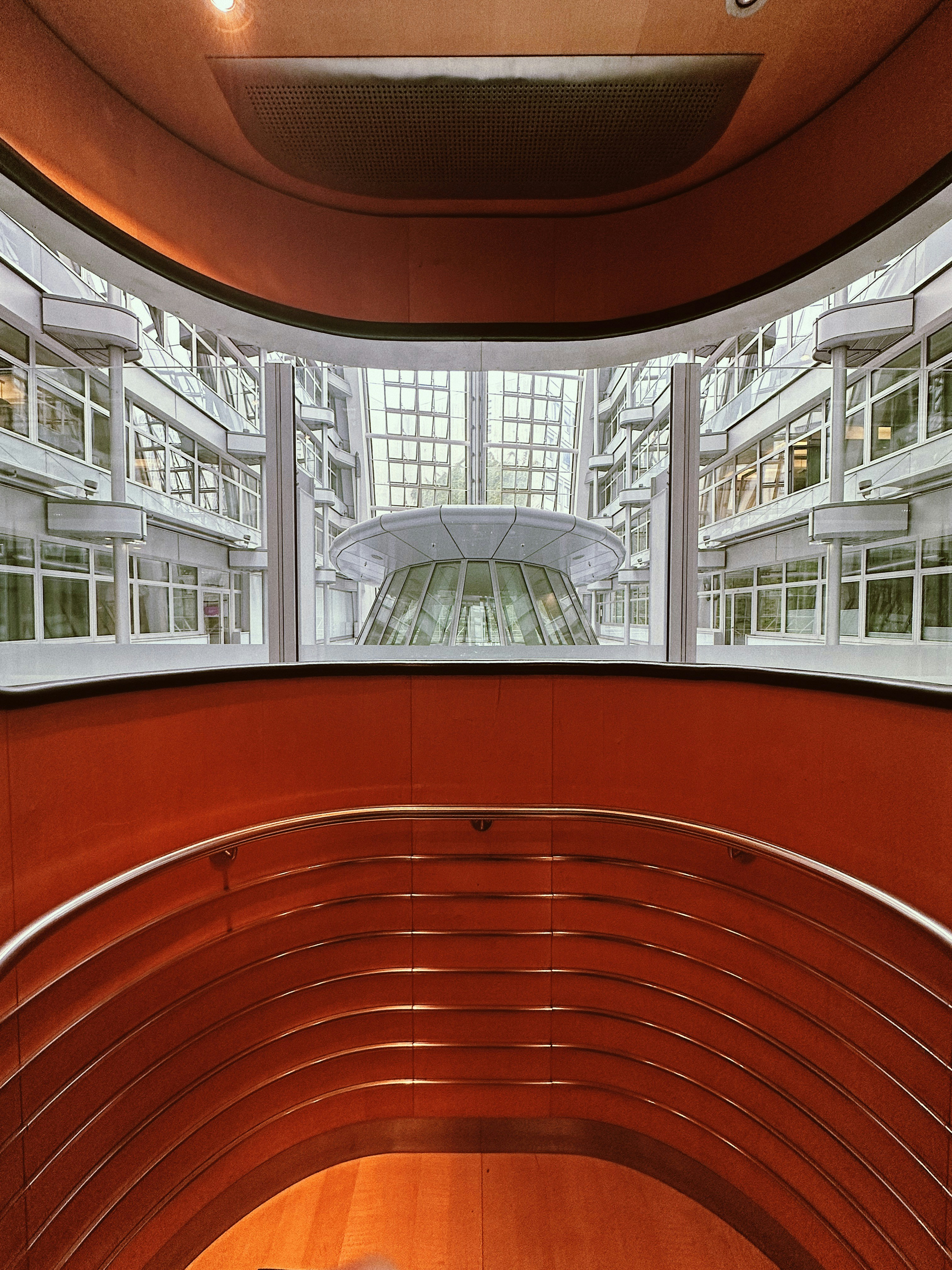 An architectural view reveals a modern indoor space with curving walls and metal railings overlooking a large glass-covered atrium. The design features a balance of metal and wood elements, highlighted by natural light from the expansive windows.