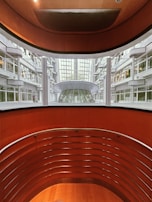 An architectural view reveals a modern indoor space with curving walls and metal railings overlooking a large glass-covered atrium. The design features a balance of metal and wood elements, highlighted by natural light from the expansive windows.