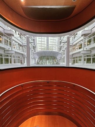 An architectural view reveals a modern indoor space with curving walls and metal railings overlooking a large glass-covered atrium. The design features a balance of metal and wood elements, highlighted by natural light from the expansive windows.