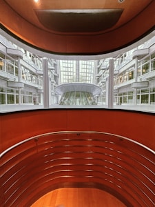 An architectural view reveals a modern indoor space with curving walls and metal railings overlooking a large glass-covered atrium. The design features a balance of metal and wood elements, highlighted by natural light from the expansive windows.