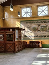 A vintage-style kiosk with wooden panels and decorative patterns is situated in a space with high ceilings. The walls are made of yellow tiles and feature large windows with cross-patterned frames. A historic poster on the wall displays images of transport systems with dates, hinting at an exhibition theme. Overhead, two hanging lights illuminate the kiosk area, creating a mix of natural and artificial lighting.