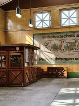 A vintage-style kiosk with wooden panels and decorative patterns is situated in a space with high ceilings. The walls are made of yellow tiles and feature large windows with cross-patterned frames. A historic poster on the wall displays images of transport systems with dates, hinting at an exhibition theme. Overhead, two hanging lights illuminate the kiosk area, creating a mix of natural and artificial lighting.