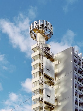A tall modern building with glass windows partially obscured by shadows, and a large vintage-style sign reading 'BERLINER' on top of the structure. The sky is blue with scattered clouds.
