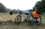 A touring bicycle rests against a wooden post, equipped with a large orange pannier on the rear wheel. The setting is a grassy field near a forest, with a distant view of hills and a partly overcast sky. Some clothing is hanging nearby, and a tent can be seen in the background, suggesting a camping scene.